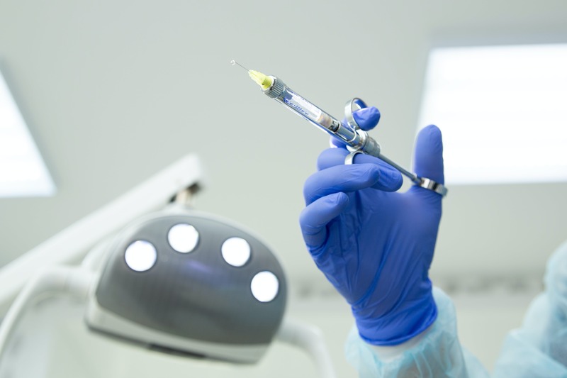 Ręka lekarza w niebieskiej rękawiczce trzyma strzykawkę do znieczulenia dentystycznego
