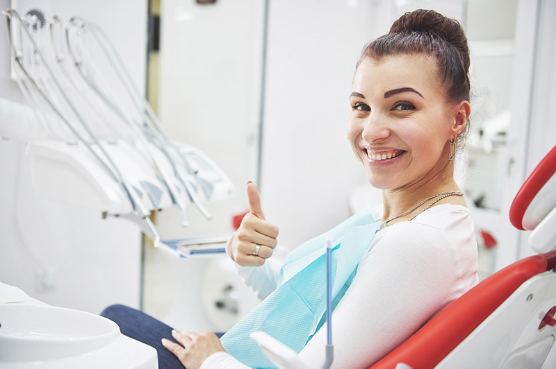 Uśmiechnięta kobieta siedzi na fotelu dentystycznym i wykonuje gest kciuka w górę i uśmiecha się